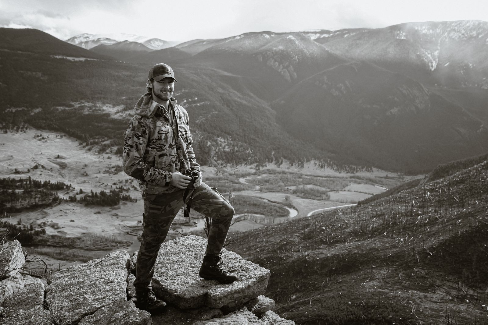 Jonah smiling on a rock outcrop above a river valley, binoculars in hand