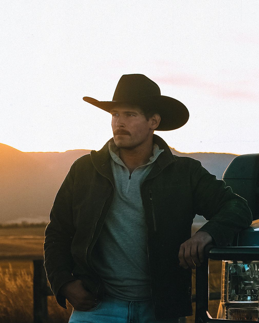 Jonah in a black felt cowboy hat and canvas jacket, leaning on a truck at sunset