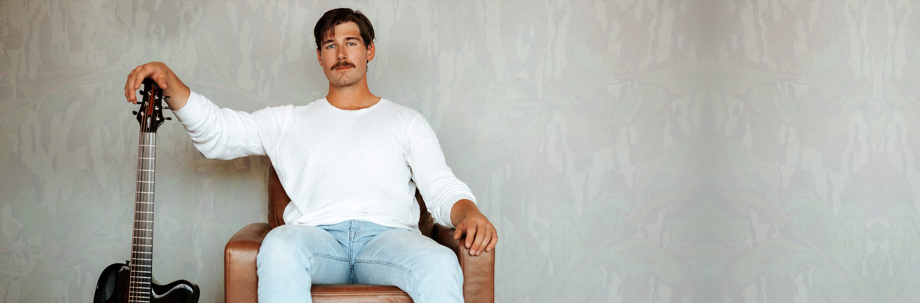 Jonah seated in a leather armchair in a white thermal and light denim, one hand resting on his guitar
