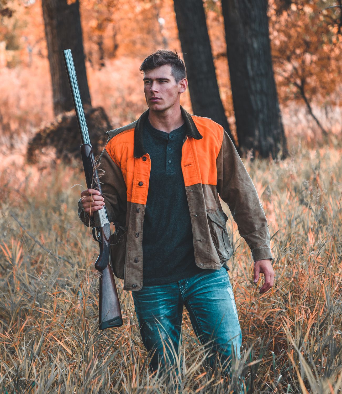Jonah walking through tall autumn grass with an over-under shotgun, wearing a blaze-orange upland jacket