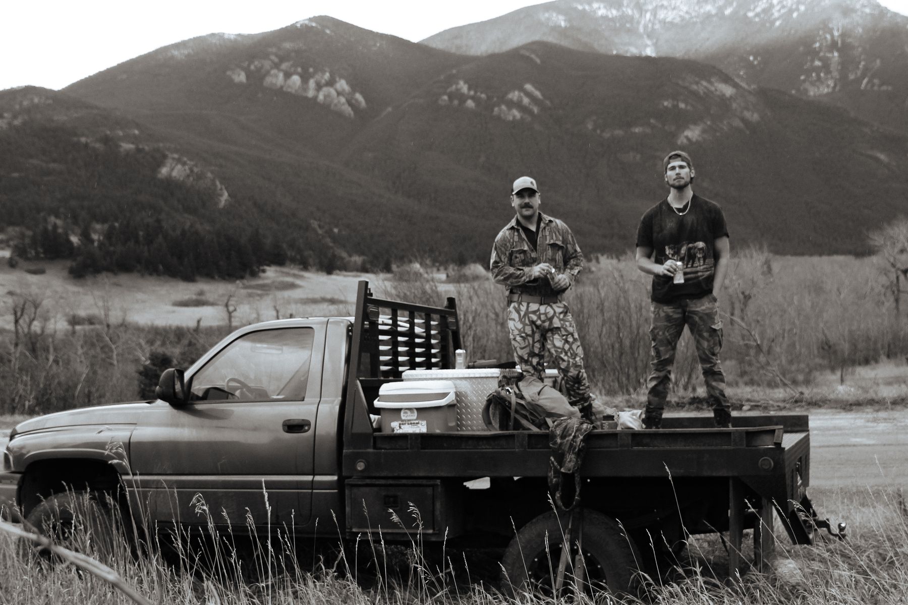 Black and white photo of Jonah and a friend in camo standing on a flatbed truck below the mountains