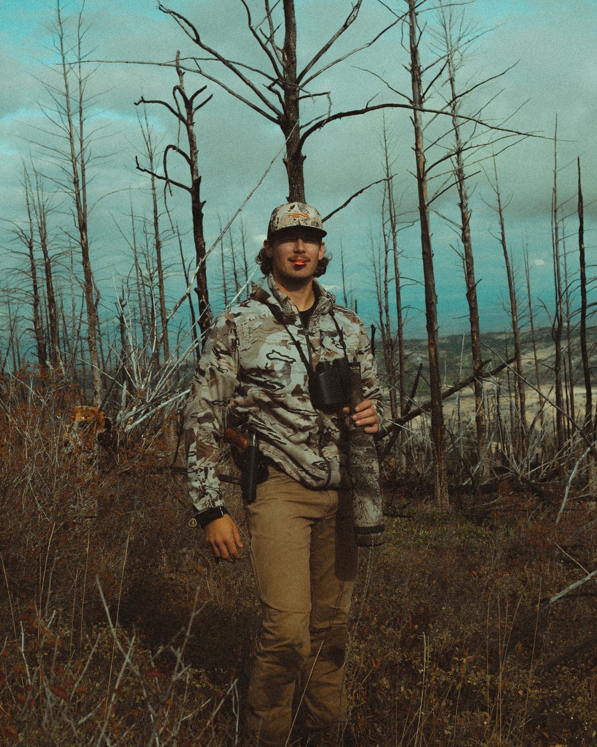 Jonah in camo among burned trees with an elk call in his mouth and a bugle tube in hand