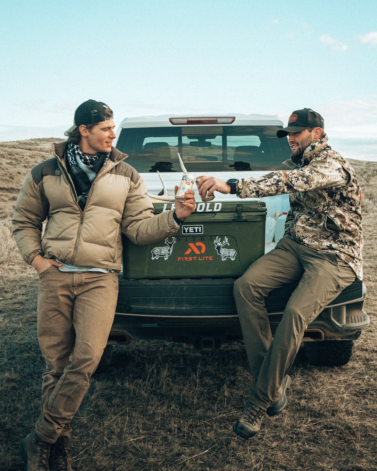 Jonah and a friend toasting at a truck tailgate with a cooler and an antler