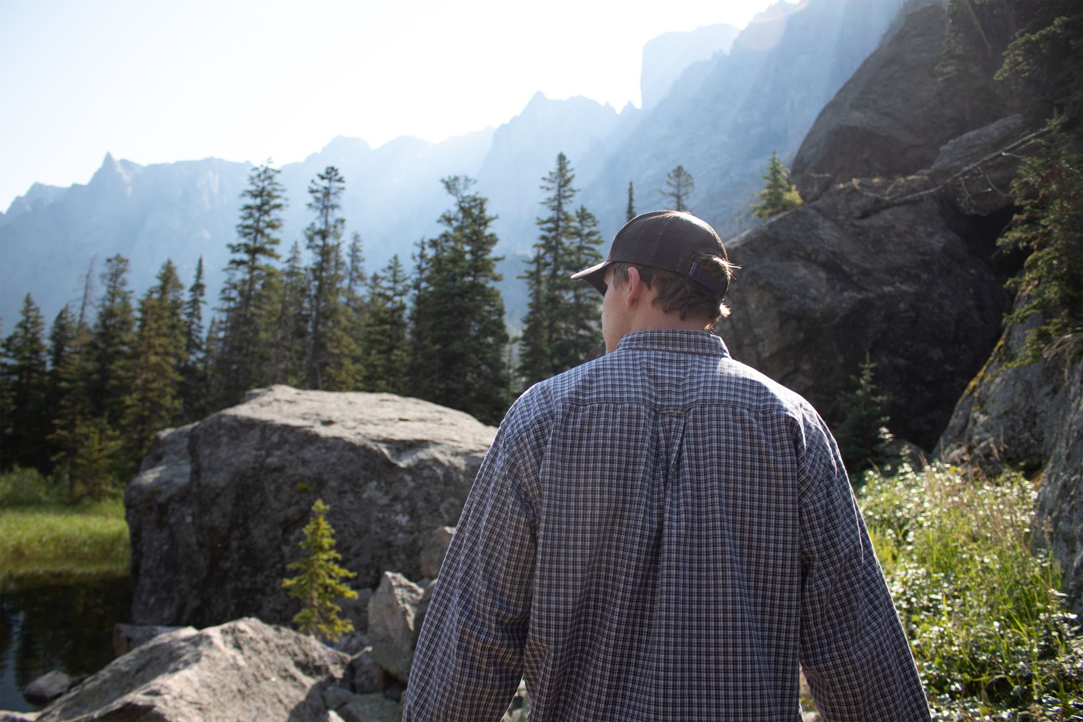 Jonah from behind in a plaid shirt and cap, walking toward sunlit granite cliffs and pines