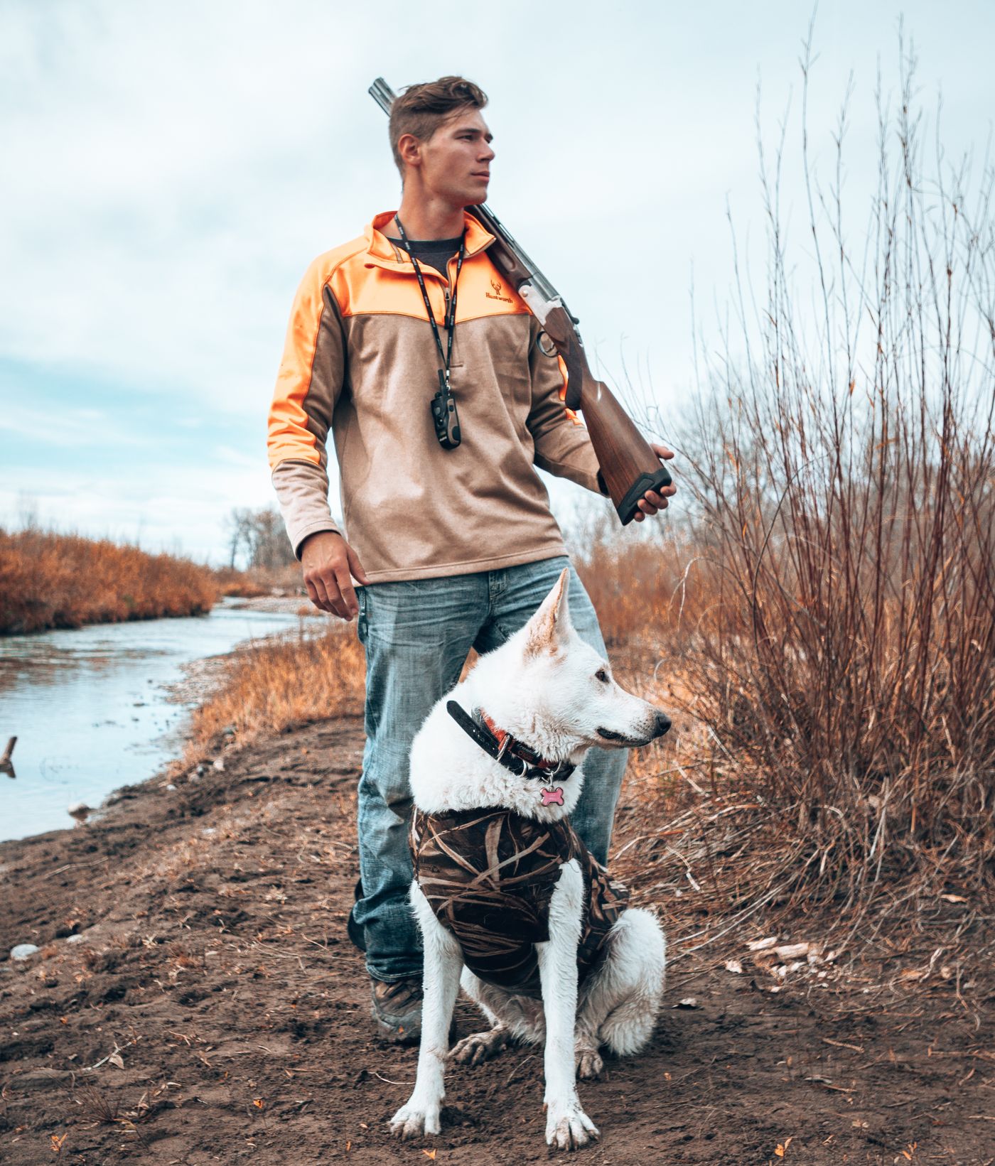 Jonah with a shotgun on his shoulder and a white German Shepherd at his feet by a river