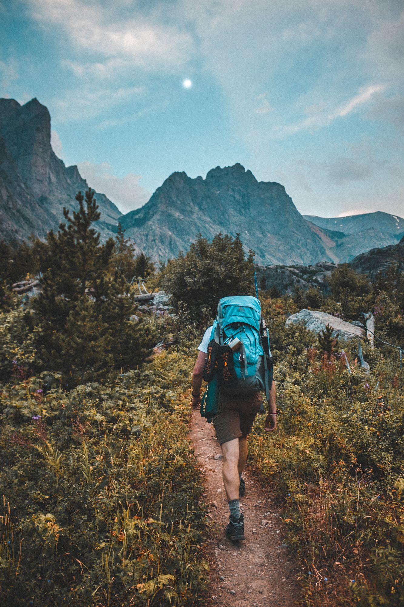 A backpacker with a blue pack walking a dirt trail toward jagged peaks